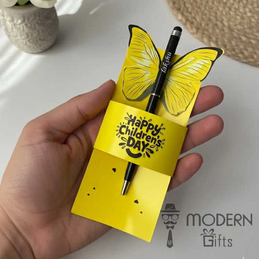 Yellow butterfly-shaped card with 'Happy Children's Day' text held by a hand, with a pen and vase in the background.