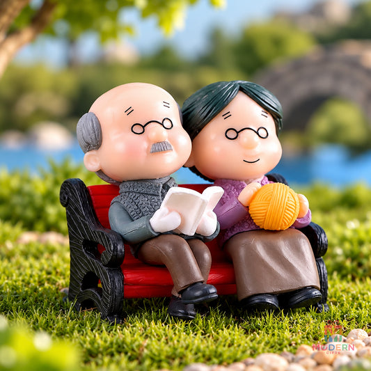 Granny Miniature Doll – Cute elderly couple miniature dolls sitting on a park bench, with the granny holding a ball of yarn and grandpa reading a book, displayed in a beautiful outdoor garden setting.
