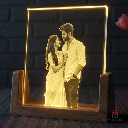 A personalized LED acrylic picture frame on a wooden stand, displaying a romantic couple portrait illuminated in warm yellow light against a dark brick wall background.